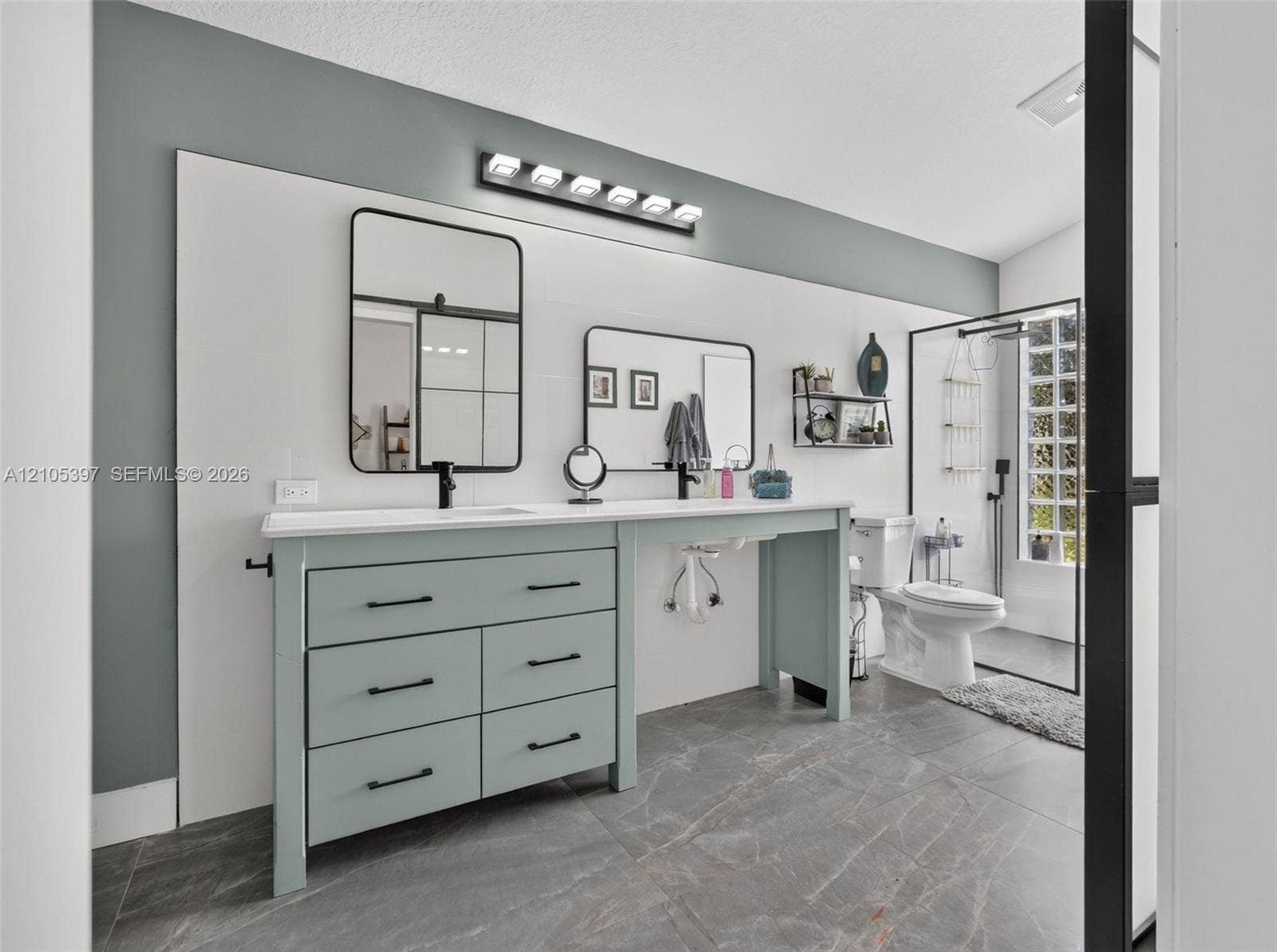 Dual-sink vanity with matte black hardware, rectangular wall-mounted mirrors, stone-finish tile flooring, integrated light fixture, and glass-enclosed shower stall ADA accessible