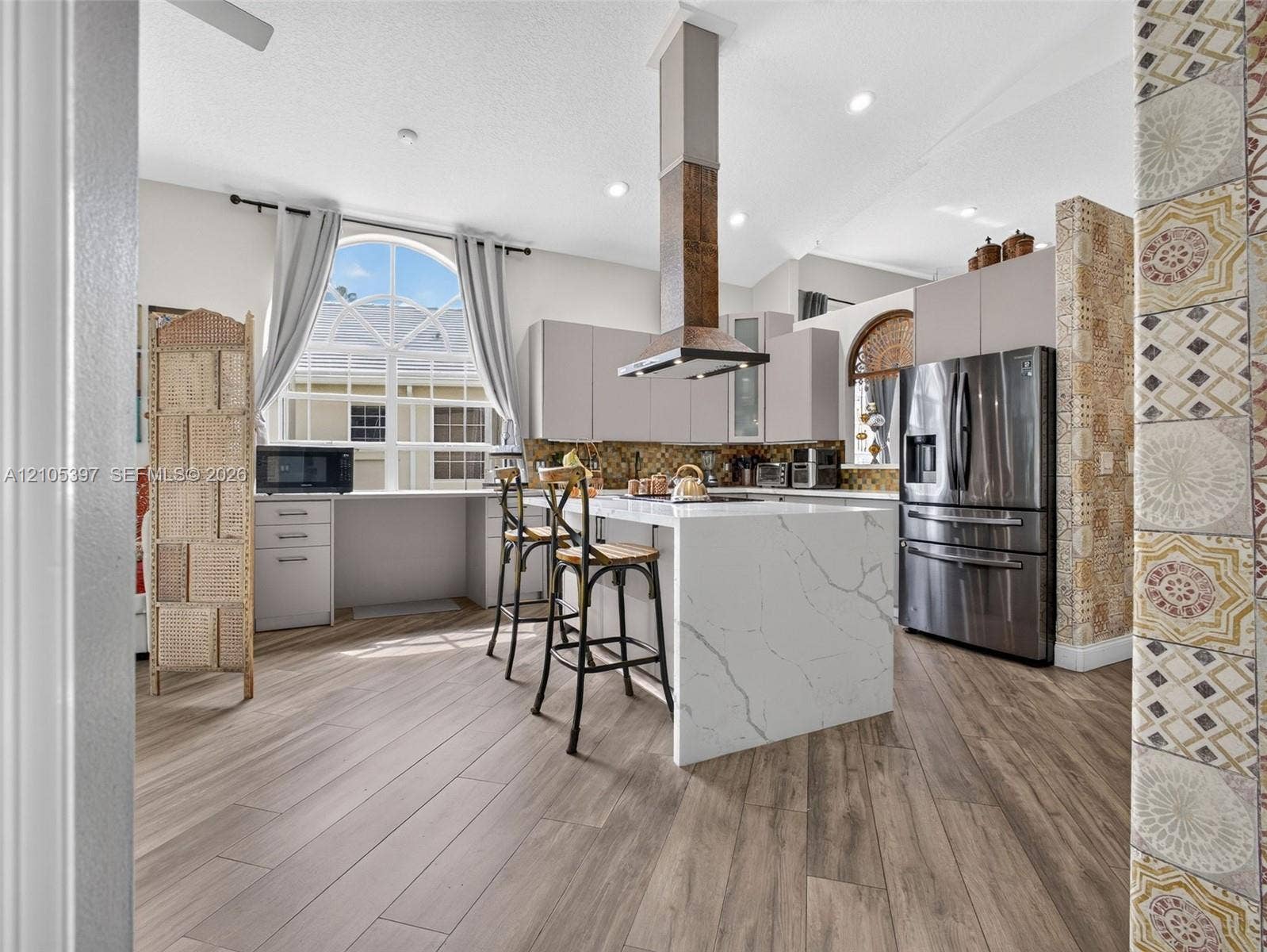 Kitchen featuring a waterfall-edge stone island, stainless steel refrigerator, tall range hood, porcelain wood-finish flooring,