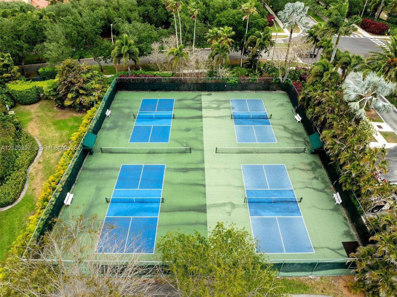 Four-court recreational facility featuring hard-surface playing areas, perimeter chain-link fencing, built-in benches, and mature professional landscaping