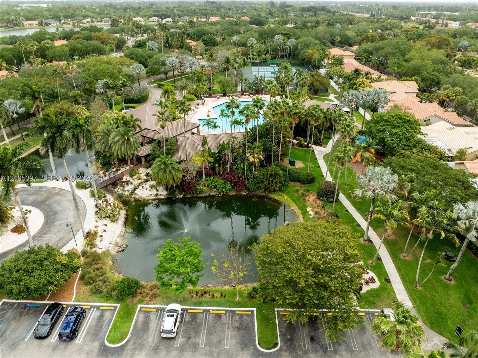 Aerial view of the community grounds featuring a central pond with a fountain, swimming pool, tennis court, and extensive landscaped walking paths