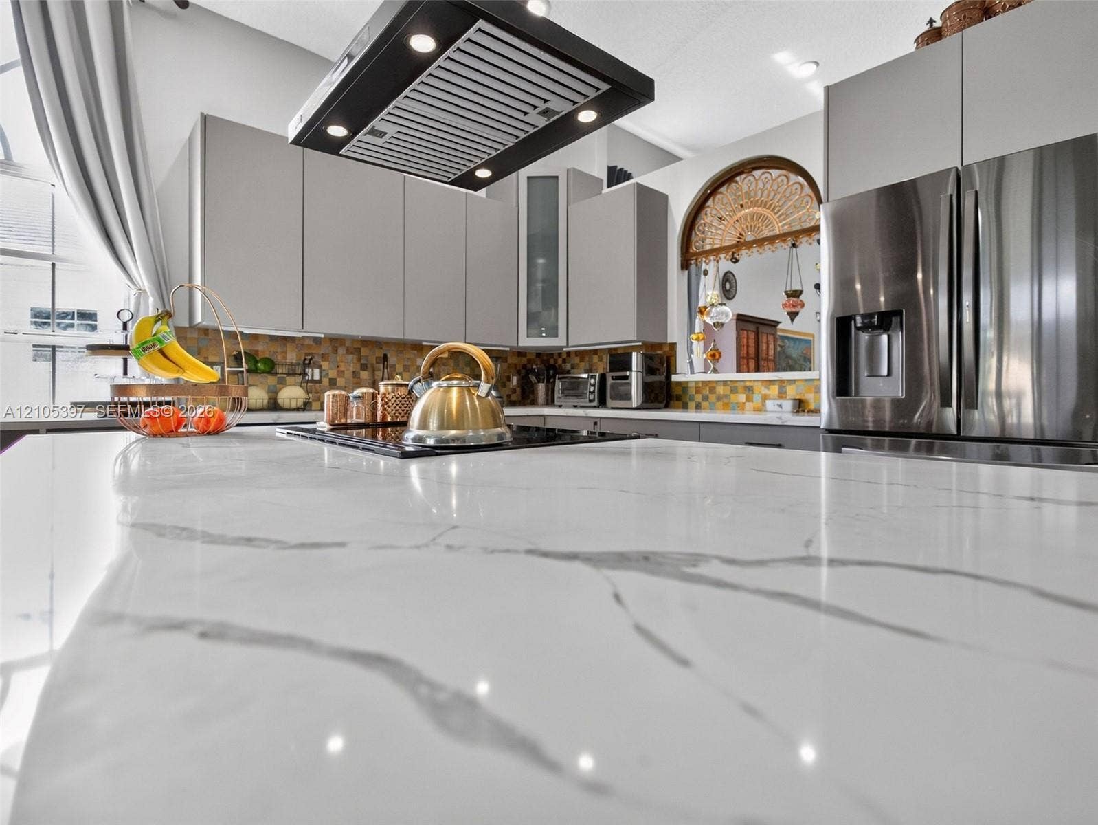 Kitchen featuring a large waterfall-edge island with stone-finish countertop, flat-panel cabinetry, stainless steel refrigerator, and built-in rectangular range hood