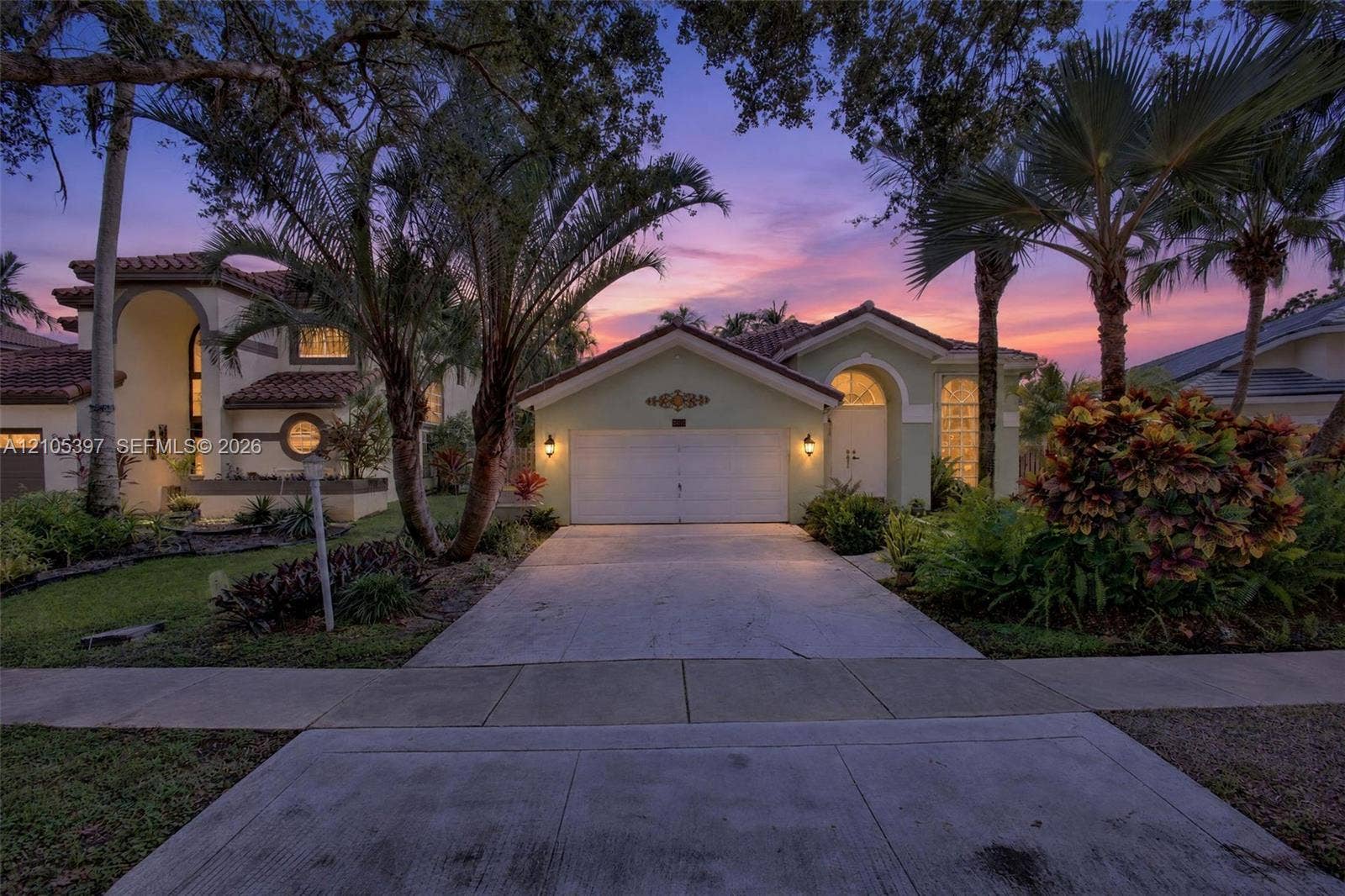 Stucco exterior featuring arched architectural detailing, a multi-car garage, concrete driveway, tiled roofing, and mature tropical landscaping