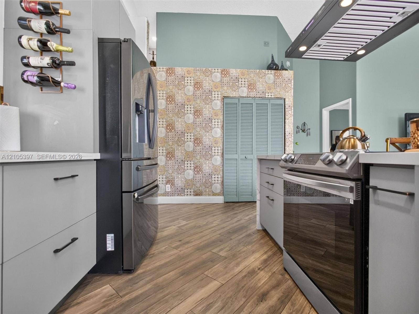 Kitchen featuring grey flat-panel cabinetry, wood-finish flooring, patterned tile accent wall, stainless steel appliances, and a mounted range hood
