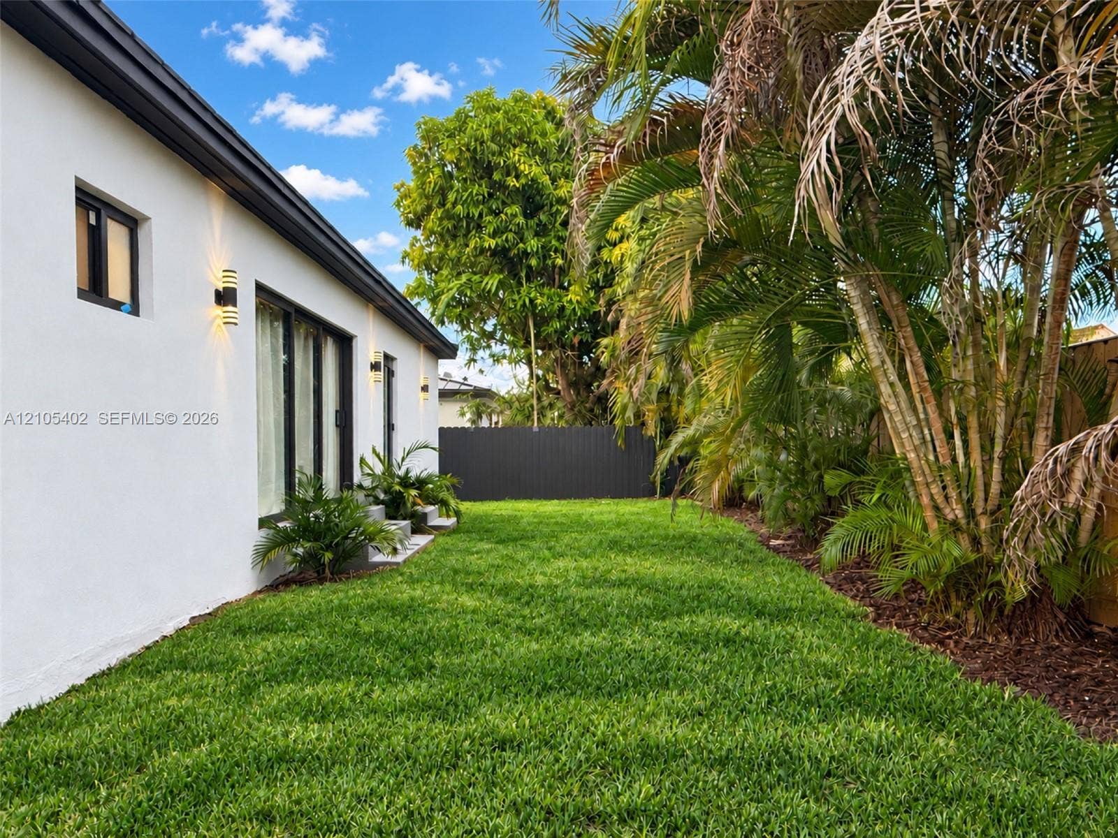 1911 SW 18th Avenue, Miami, FL 33145 Exterior featuring white stucco siding, black-framed glass sliding doors, dark metal wall-mounted exterior lighting, manicured lawn, and mature palm trees enclosed by a charcoal-colored privacy fence