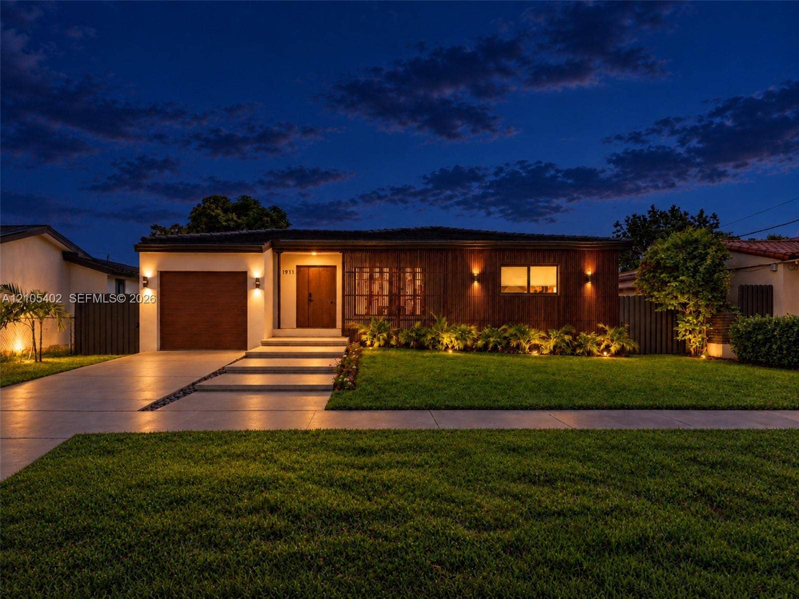 1911 SW 18th Avenue, Miami, FL 33145 Modern single-story exterior featuring a wood-panel faade, integrated wall sconce lighting, solid wood front door, horizontal-slat garage door, and concrete entry steps