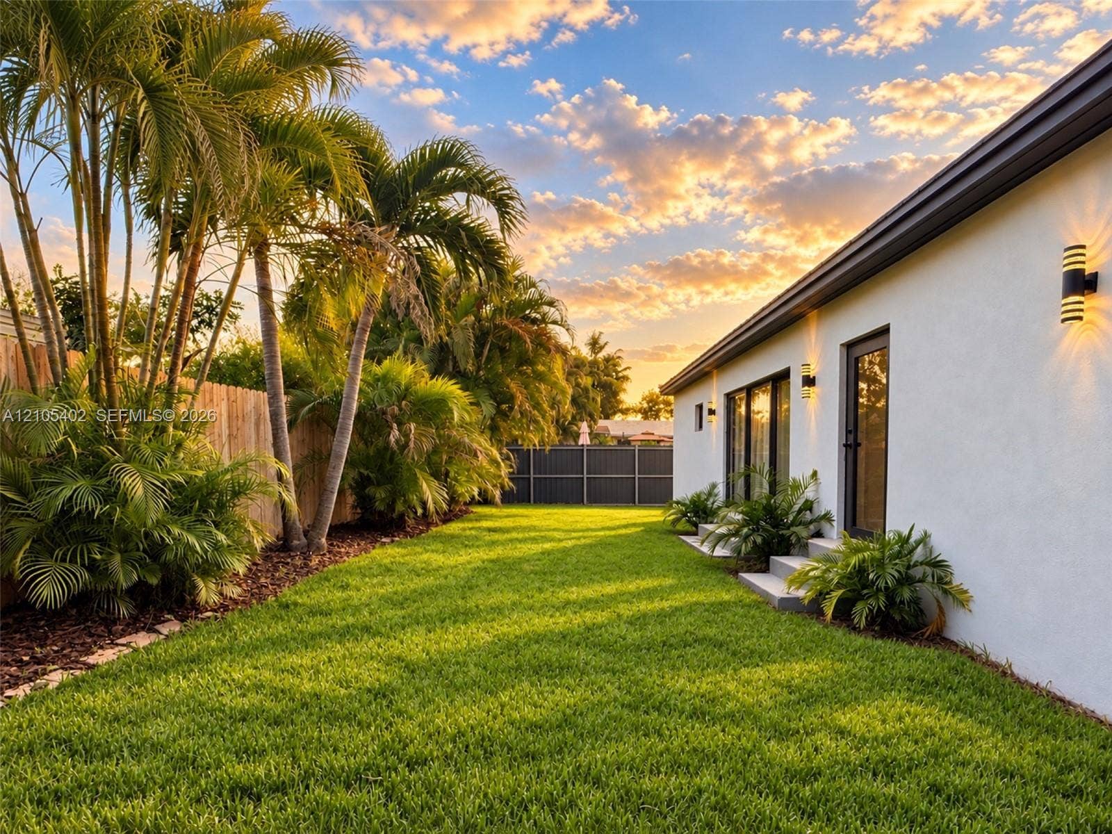 1911 SW 18th Avenue, Miami, FL 33145 Expansive backyard featuring a manicured lawn, mature palm trees, exterior wall-mounted lighting, and privacy fencing