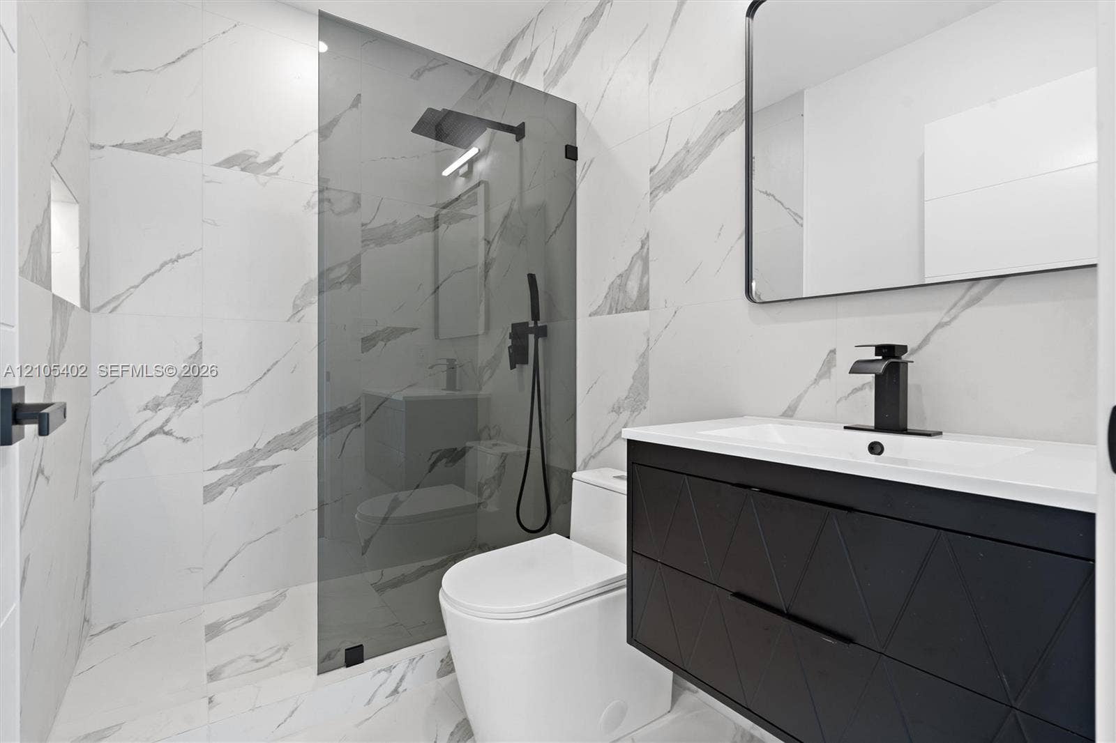 1911 SW 18th Avenue, Miami, FL 33145 Modern bathroom featuring large-format wall and floor tile with a veined pattern, walk-in shower with glass panel, matte black plumbing fixtures, and a textured floating vanity with integrated sink