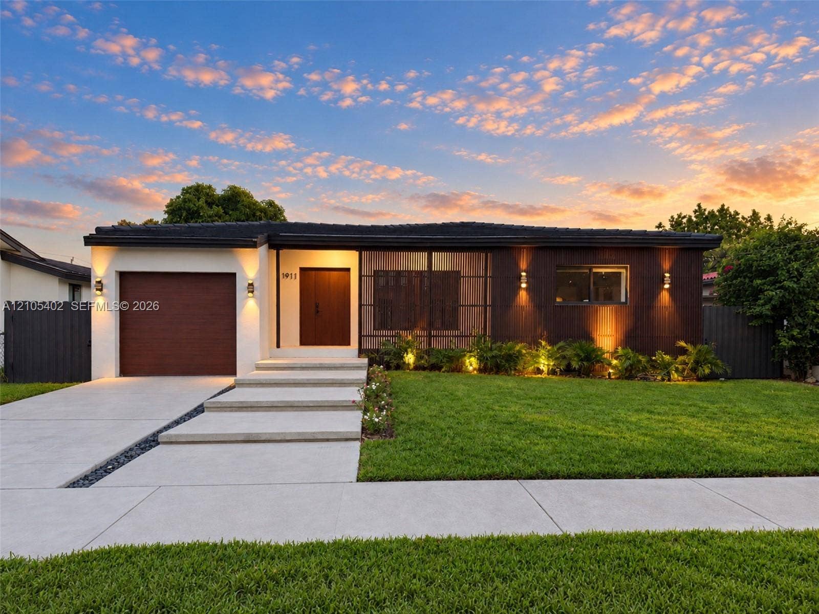 1911 SW 18th Avenue, Miami, FL 33145 Single-story residence featuring a mid-century modern aesthetic, wood-paneled exterior accents, a concrete driveway with stone drainage detail, a stepped concrete entryway, and wall-mounted exterior lighting