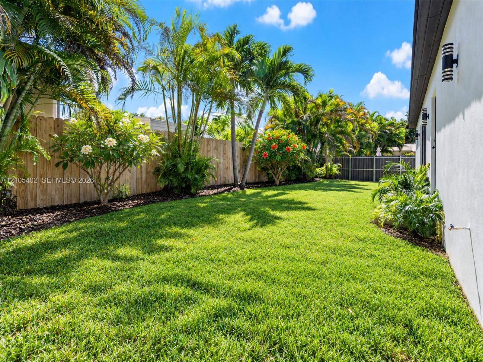 1911 SW 18th Avenue, Miami, FL 33145 Landscaped backyard featuring a manicured lawn, wood-panel fencing, mature palm trees, and exterior wall-mounted light fixtures