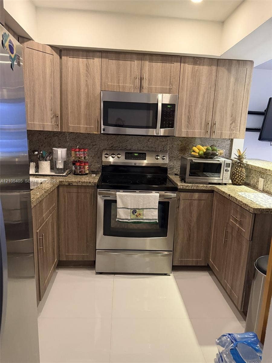 17570 Atlantic Boulevard, #119, Sunny Isles Beach, FL 33160 U-shaped kitchen featuring wood-finish cabinetry, stainless steel appliances, speckled countertops, and a full-height backsplash