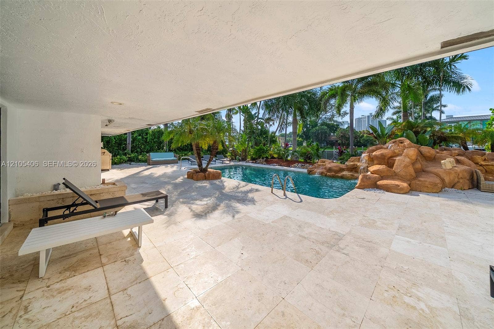 Expansive covered patio featuring light-toned tile flooring, directly accessing a custom pool with a rock waterfall feature and integrated steps