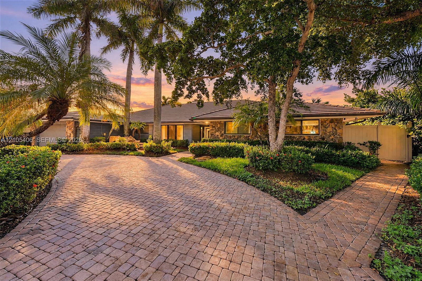 Expansive paver driveway with split path, lush landscaping, and mature palm trees framing a stone-clad exterior