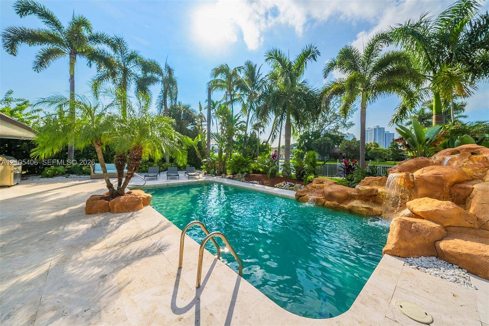 Resort-style pool area featuring a natural rock waterfall, integrated palm trees, light-toned paver decking, and lush tropical landscaping