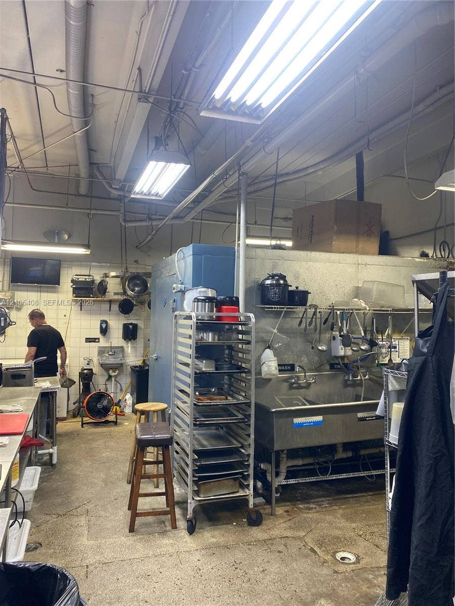 Commercial-style kitchen featuring multiple fluorescent light fixtures, exposed ductwork, a stainless steel sink with mounted shelving, and a large reach-in refrigerator unit