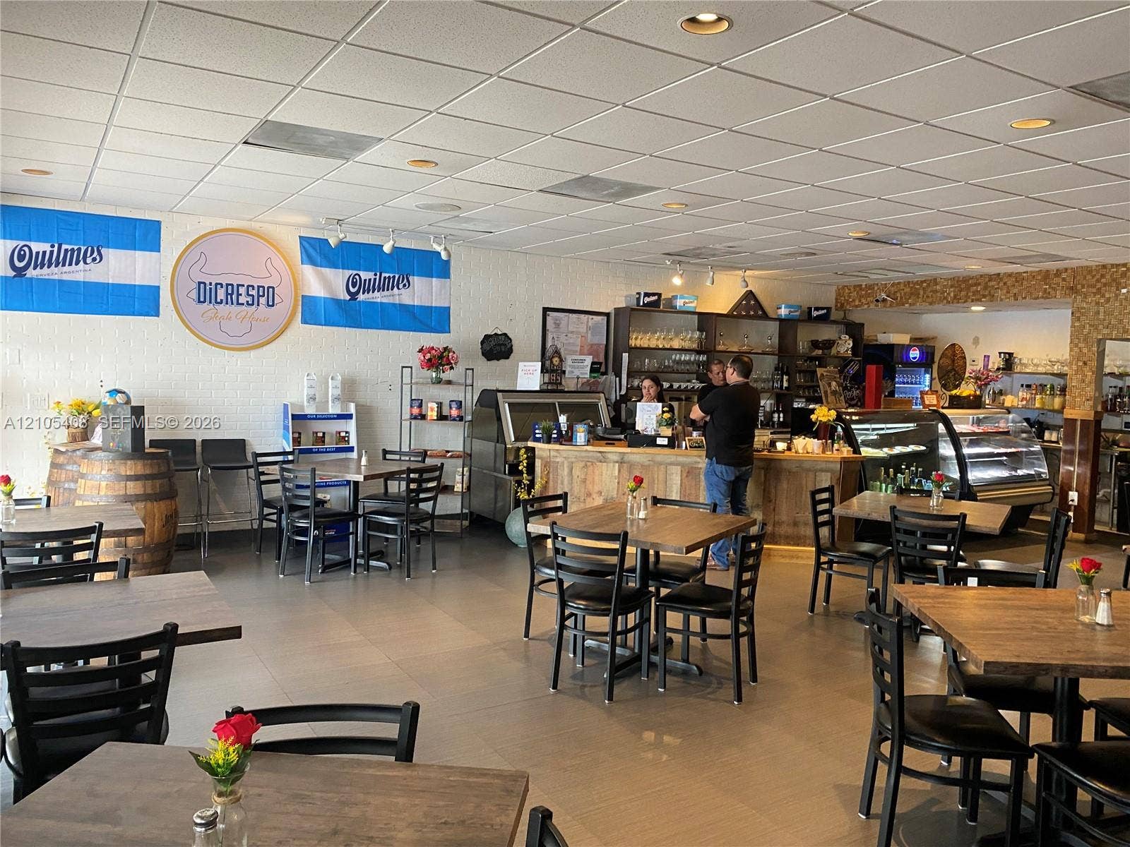 Recessed lighting illuminates the open retail space with a drop ceiling, featuring a service counter with wood-finish paneling, a display refrigeration unit, and a tiled accent wall