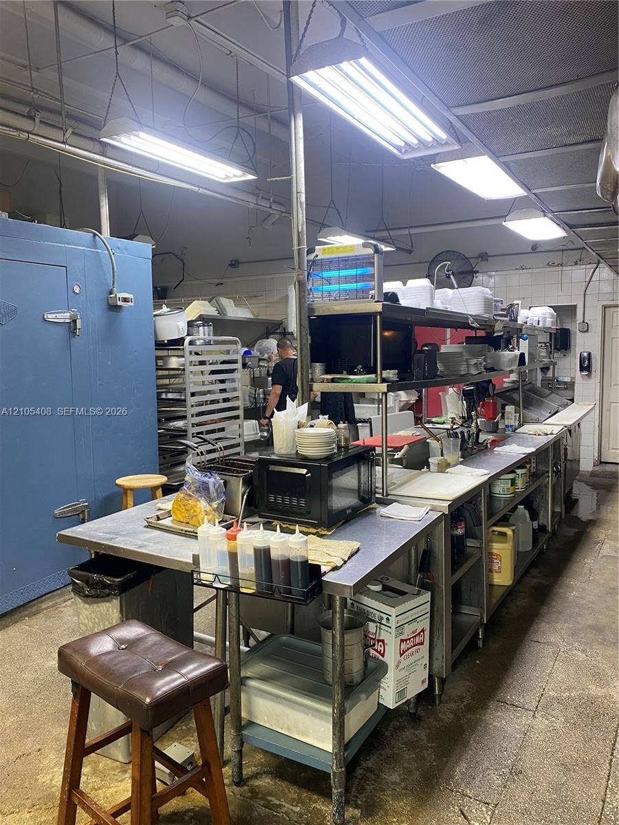 Commercial kitchen with extensive stainless steel countertops, multiple fluorescent lighting fixtures, and a large blue walk-in refrigeration unit