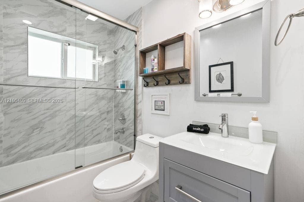 Built-in tub with glass enclosure, large format wall tile, single vanity with integrated sink, wall-mounted mirror, and wall-mounted light fixture