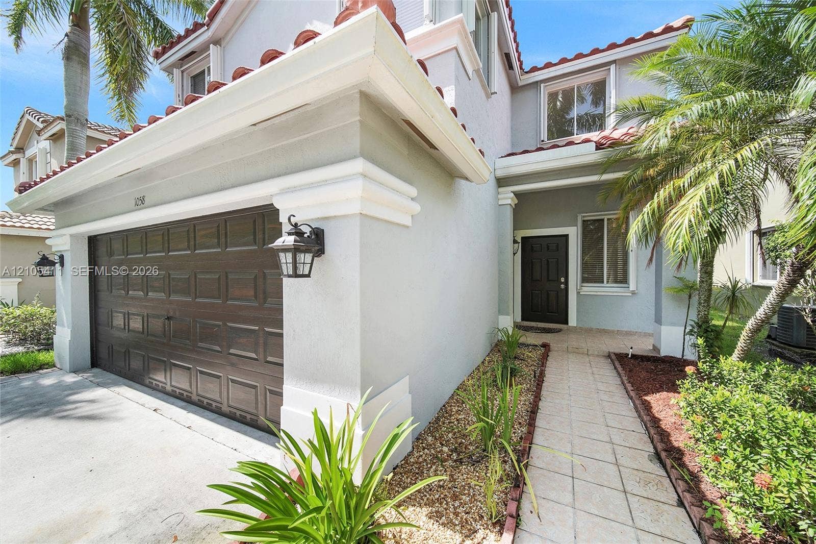 Tiled roof with white gutters, wood-paneled garage door, exterior wall sconces, tiled entryway path, and mature landscaping