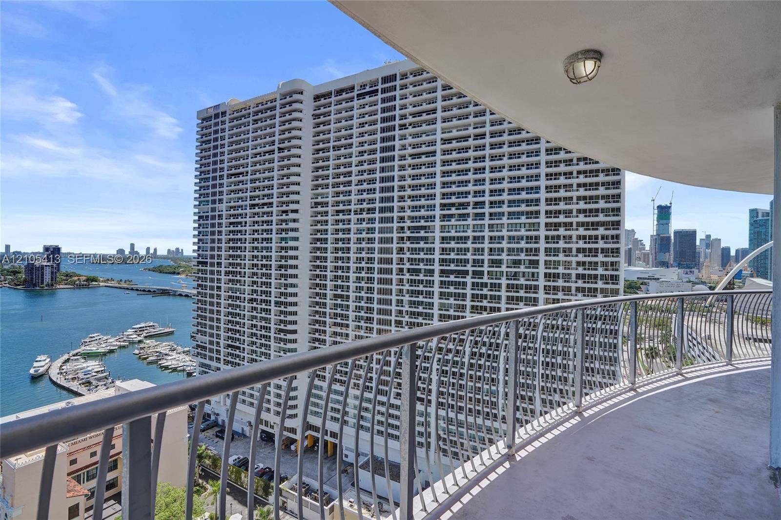 Curved balcony with metal railing, waterfront scenery, marina with multiple boats, distant cityscape, and prominent high-rise buildings