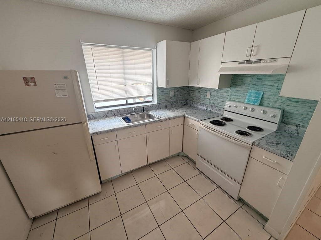 White cabinetry with chrome hardware, coordinating white appliances, a stainless steel sink, and light-toned tile flooring