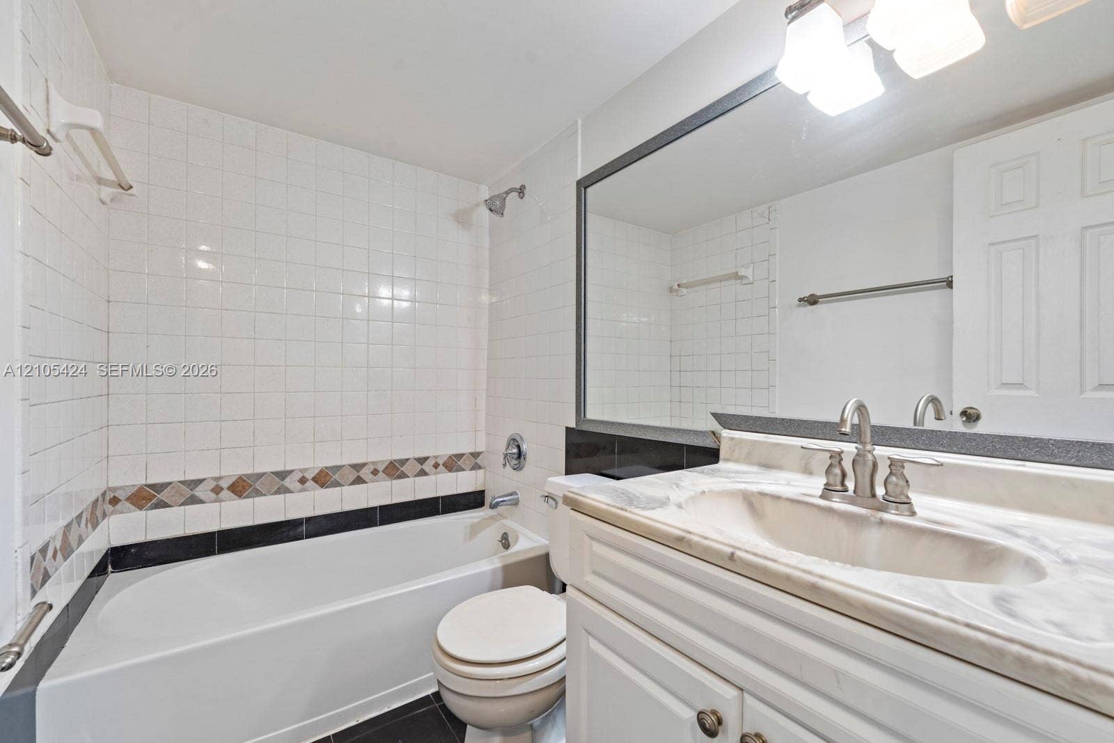 White tile shower surround with decorative accent band, tub with shower fixture, and a vanity with a white countertop and integrated sink