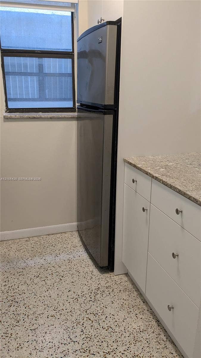Modern kitchen featuring stainless steel refrigerator with top and bottom freezer configuration, natural stone countertops, white cabinetry with brushed metal hardware, and polished concrete flooring