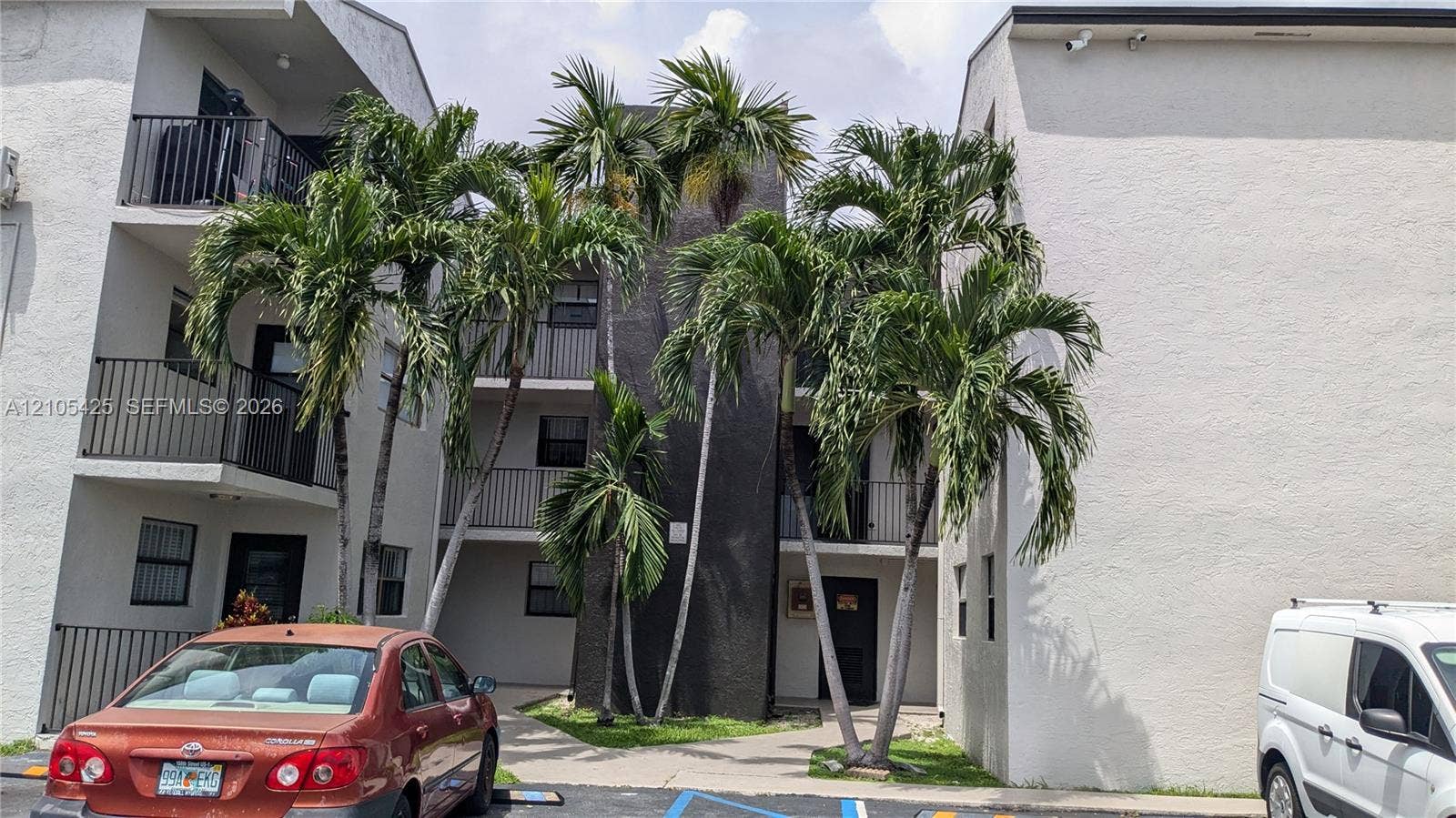 Multi-story building featuring balconies with black metal railings, textured light-colored stucco exterior, and mature palm trees