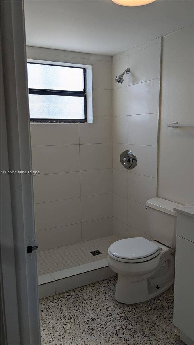 Walk-in shower with tiled walls and a window, complemented by a white toilet and a vanity with a white countertop