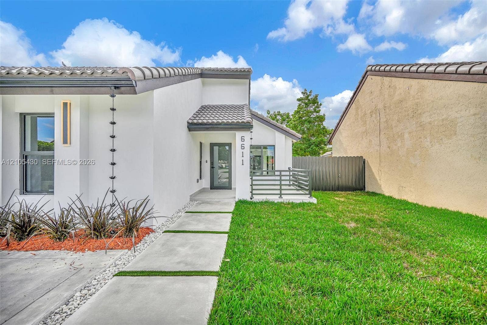 Exterior featuring smooth white stucco finish, concrete paver walkway with turf inlay, tile roof, vertical metal rain chain, and front patio with horizontal railing