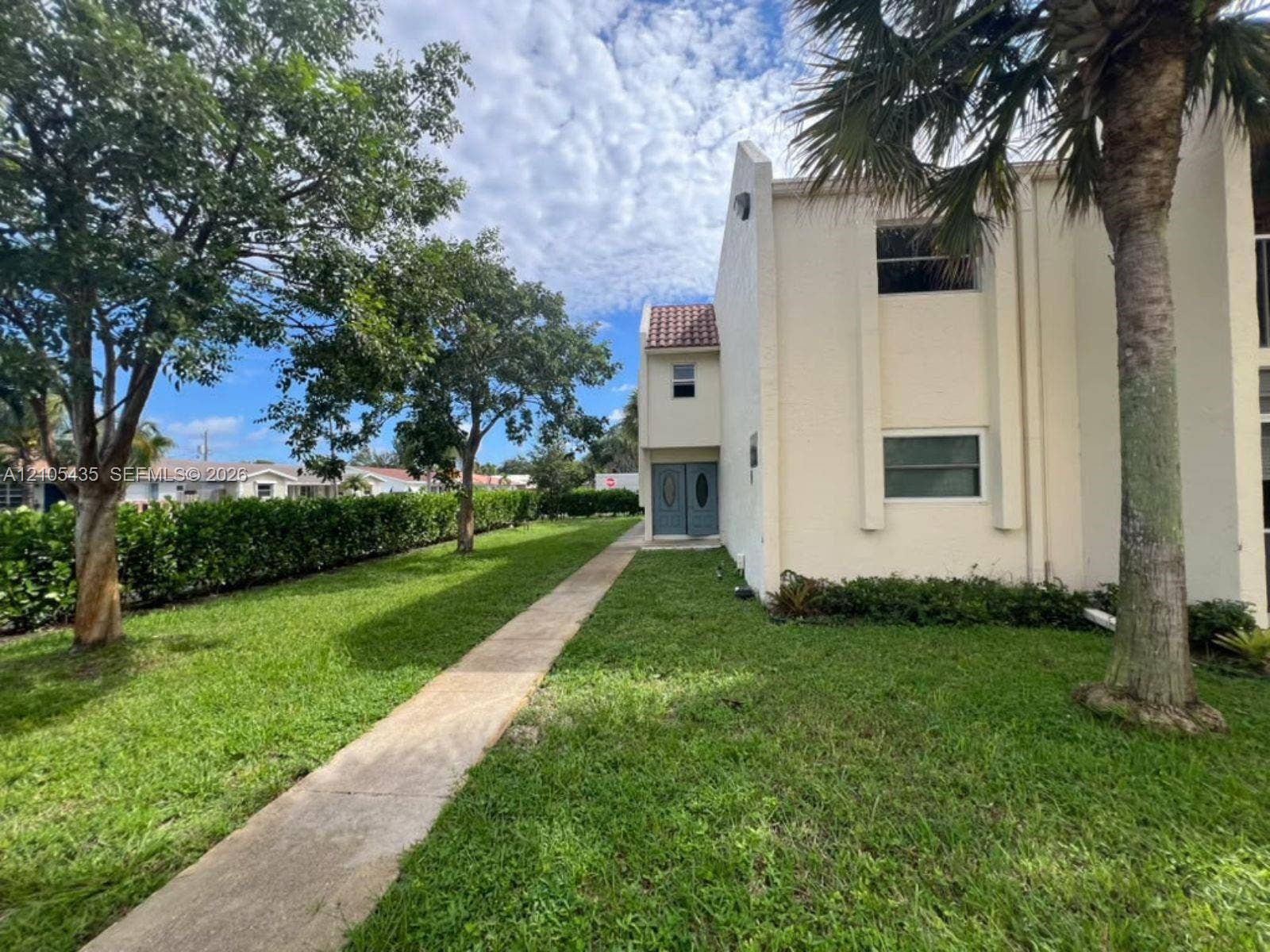 1428 SE 4th Avenue, #159, Deerfield Beach, FL 33441 Exterior facade featuring light-toned masonry, a concrete paver walkway, double-door entryway, and mature palm trees situated on a grassy lawn