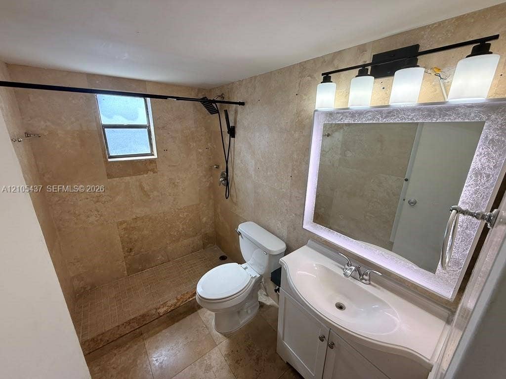 Walk-in shower with natural stone-finish wall and floor tile, integrated window, single vanity with white cabinetry and integrated sink, and an illuminated mirror