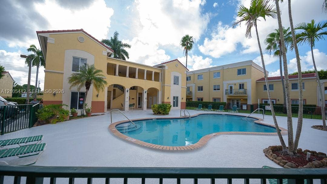 Resort-style pool with a white deck, surrounded by mature palm trees and Mediterranean-inspired architecture featuring arched walkways and tiled roofs