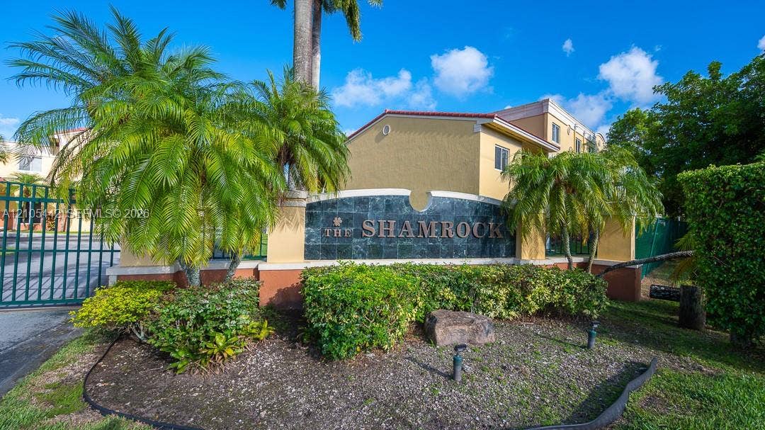 Gated community entrance with a decorative sign, lush landscaping, and mature palm trees