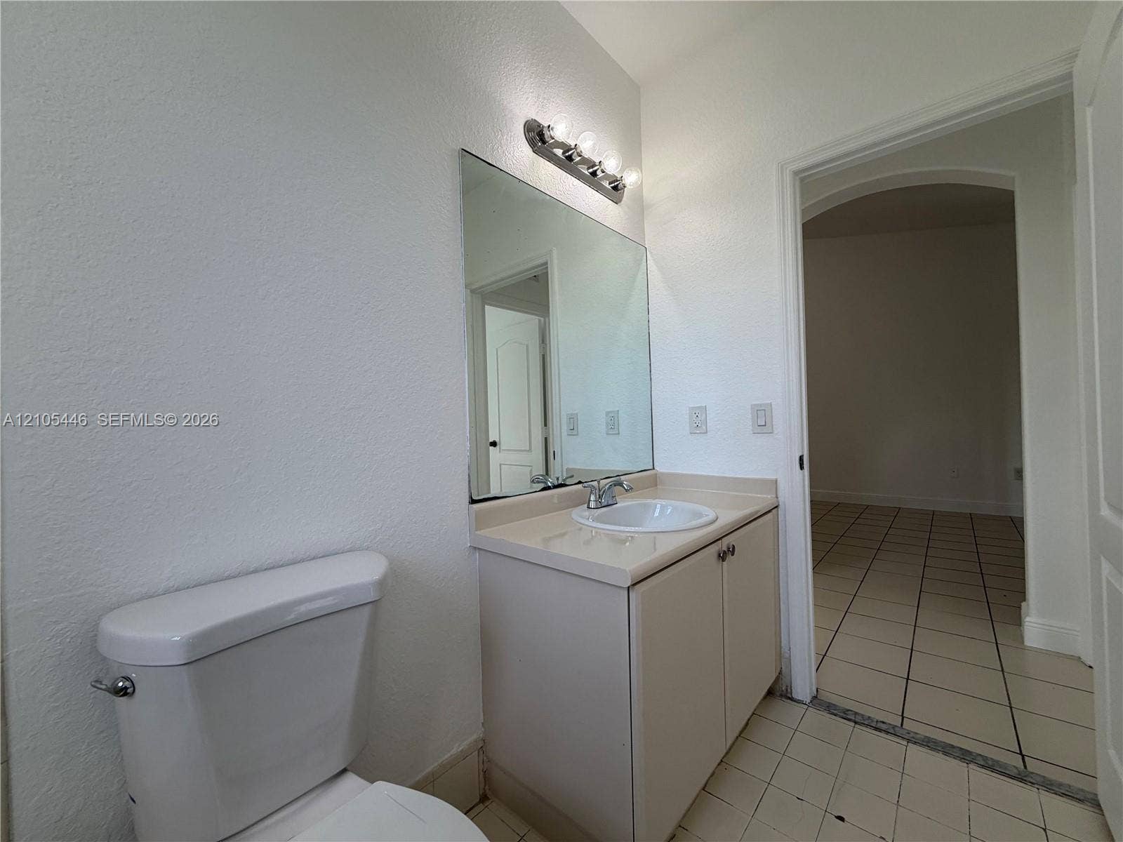 White toilet with chrome flush lever, vanity with integrated sink and chrome faucet, vanity mirror with overhead light fixture, and tile flooring