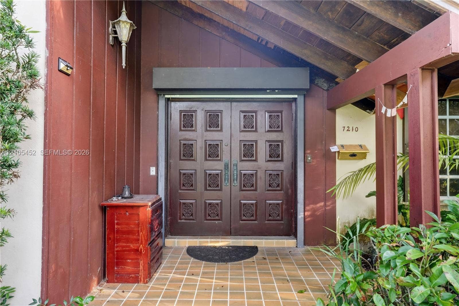 Double entry doors with detailed geometric paneling, wood-finish vertical siding, exposed beam ceiling, and ceramic tile porch