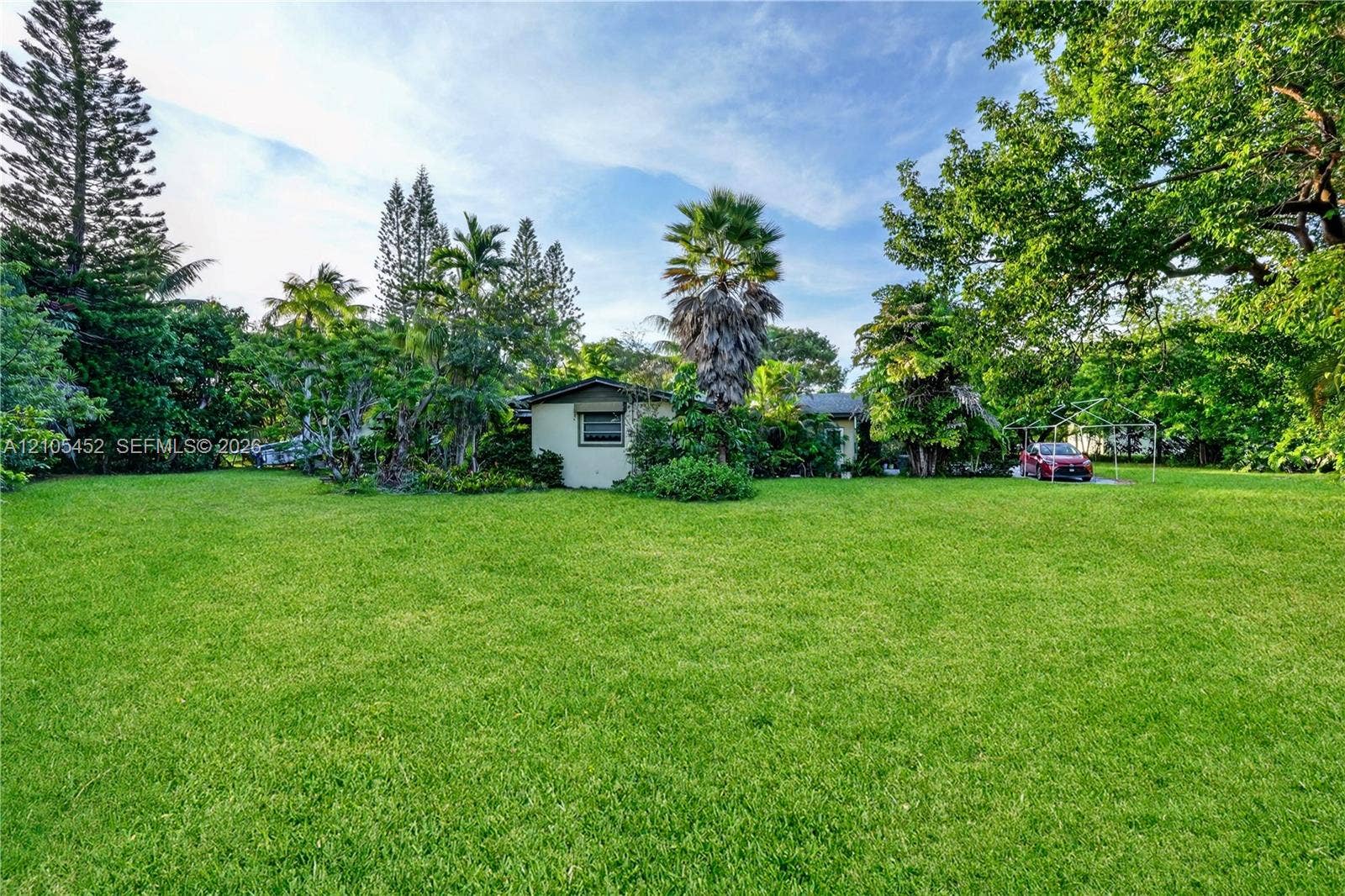 Expansive lawn featuring mature trees, diverse tropical landscaping, and a detached carport structure