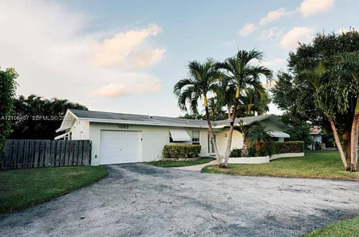 1083 SW 25th Avenue, Boynton Beach, FL 33426 Exterior facade featuring a single-car garage, white stucco siding, metal window awnings, and mature palm trees within the landscaped front lawn