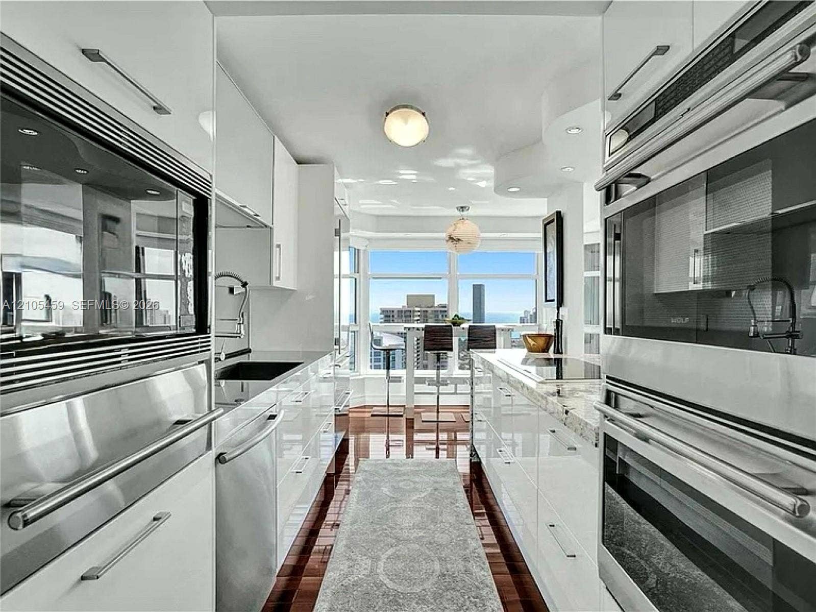 Galley kitchen featuring hardwood flooring, white high-gloss cabinetry, integrated stainless steel appliances, and stone countertops