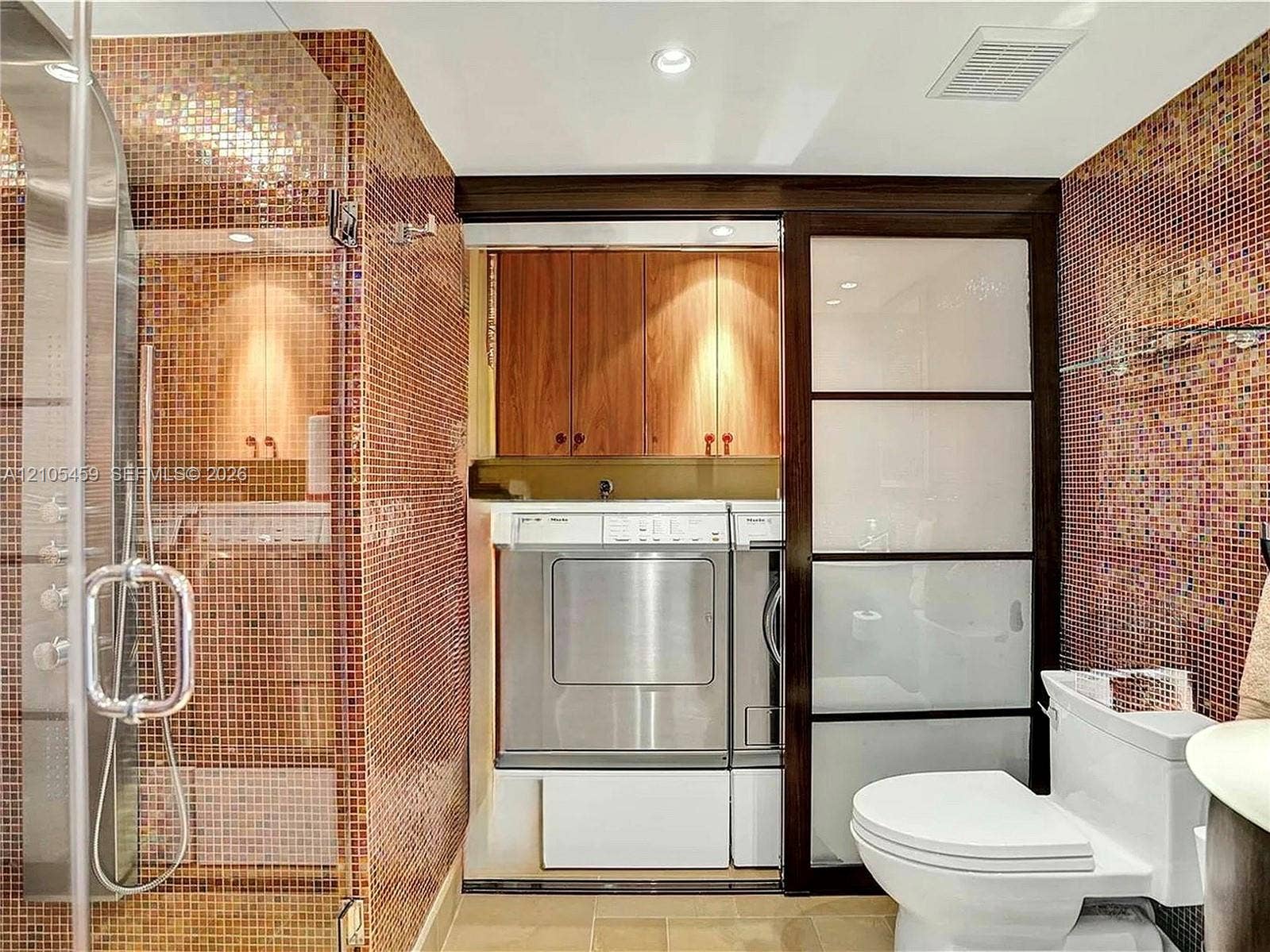 Full-height mosaic tile, glass-enclosed shower, built-in cabinetry, modern sliding door, and stackable laundry appliances