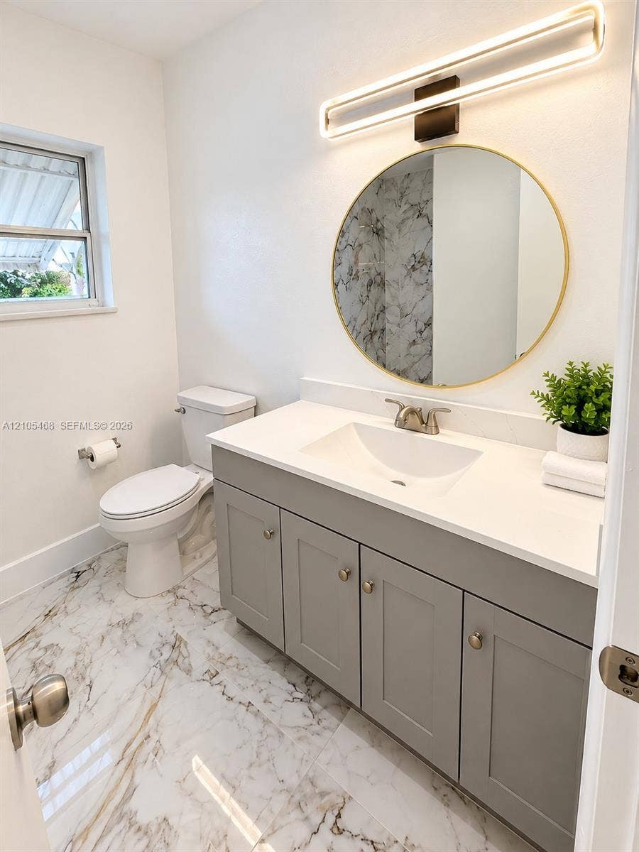 5300 Washington Street, #I201, Hollywood, FL 33021 Vanity with a white countertop and integrated sink, gray cabinetry with brass-finish hardware, a circular mirror, and a wall-mounted linear light fixture