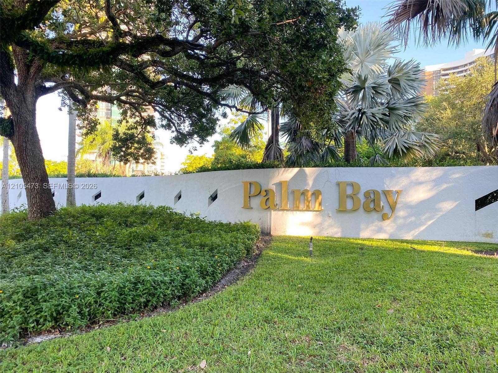 770 NE 69th Street, #6F, Miami, FL 33138 White stucco masonry wall with decorative diamond-shaped cutouts, gold-toned dimensional lettering, mature oak tree canopy, and manicured landscaping