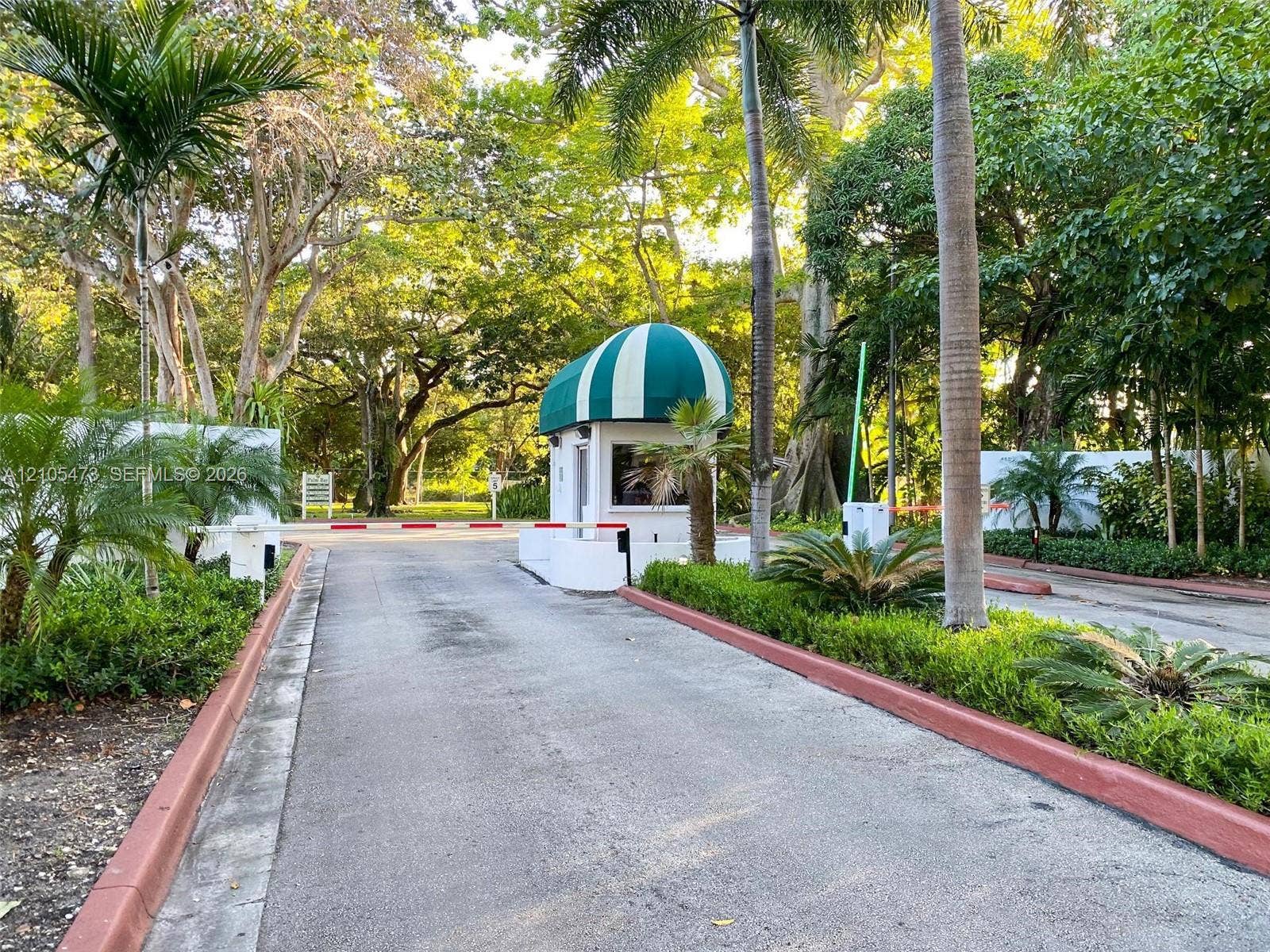 770 NE 69th Street, #6F, Miami, FL 33138 Gated entrance featuring a guardhouse with a green and white striped awning, controlled access barrier, and mature tropical landscaping