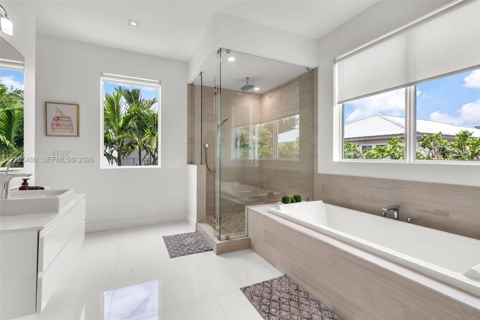 Spacious bathroom featuring white floor tile, a soaking tub with wood-finish surround, a glass-enclosed shower, dual windows, and a floating vanity with dual sinks