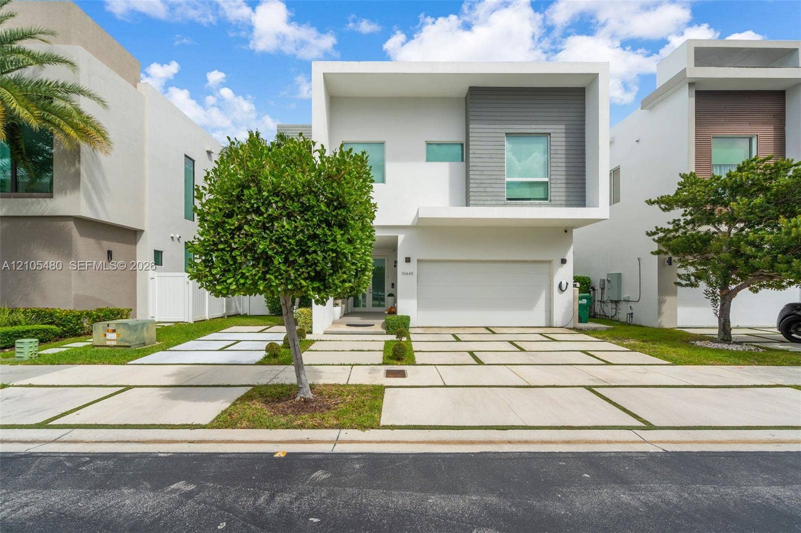 Modern architectural facade with a white and gray exterior, integrated garage, geometric driveway pavers, and established landscaping