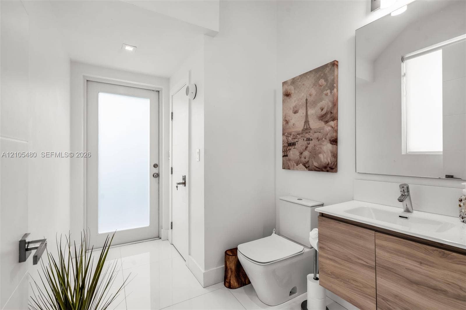 White tile flooring, contemporary vanity with wood-finish cabinetry, rectangular under-mount sink, chrome faucet, and a wall-mounted mirror with integrated lighting