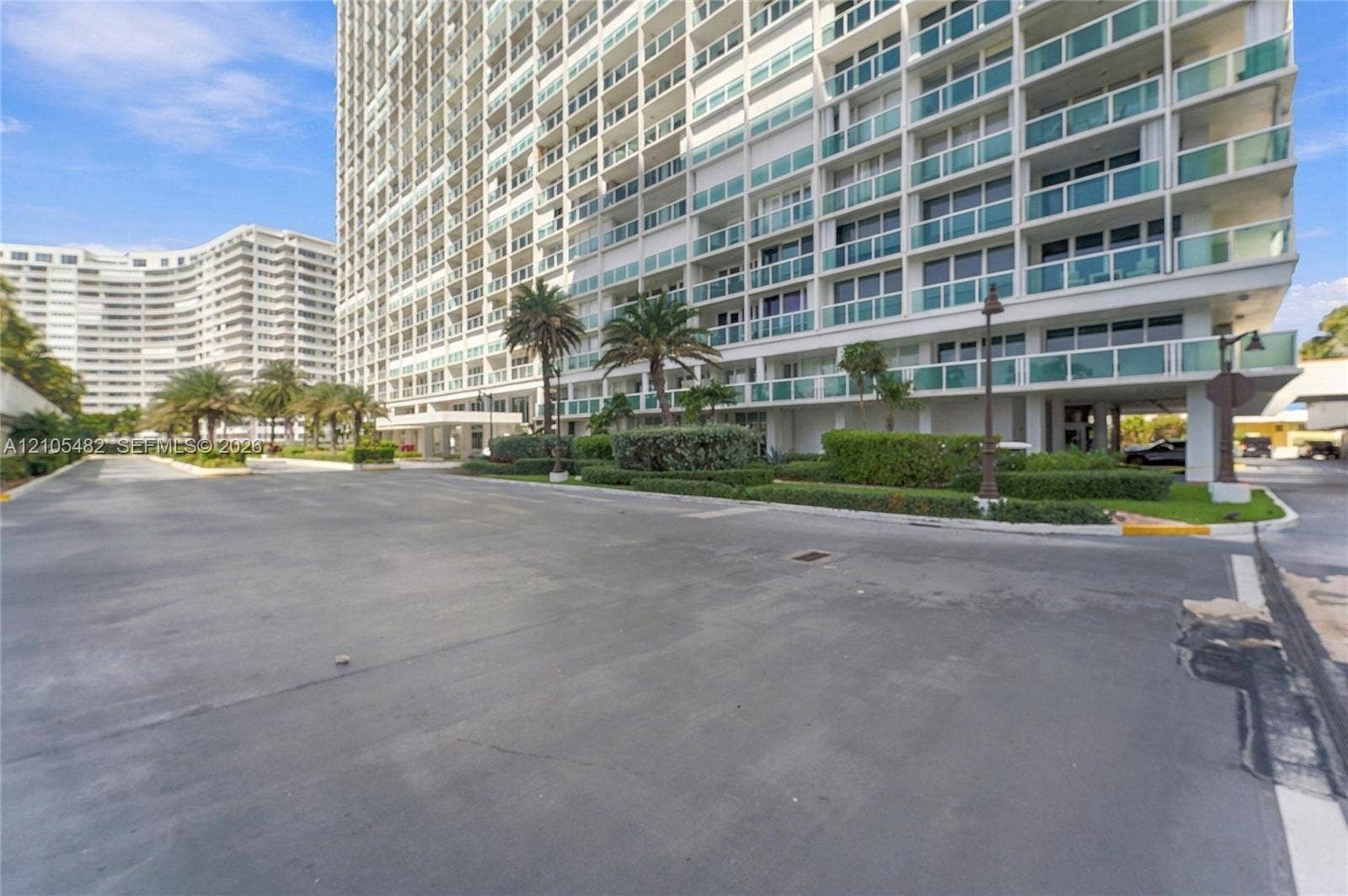 2100 S Ocean Lane, #808, Fort Lauderdale, FL 33316 High-rise building exterior featuring glass balcony railings, white structural columns, paved driveway, and mature palm tree landscaping