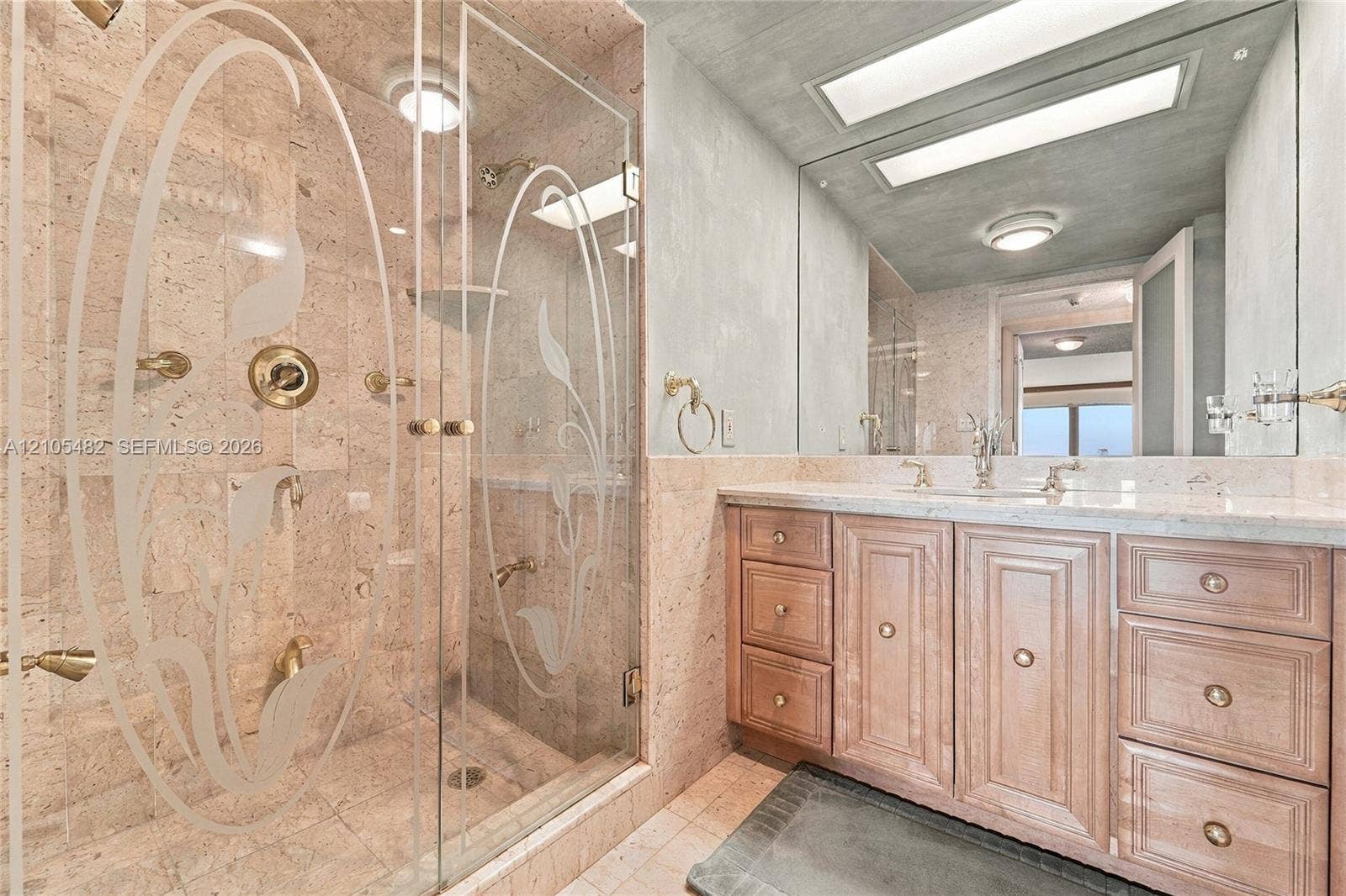 2100 S Ocean Lane, #808, Fort Lauderdale, FL 33316 Bathroom featuring a walk-in shower with etched glass doors, stone wall cladding, gold-tone fixtures, wood-finish cabinetry, and an expansive wall-to-wall mirror