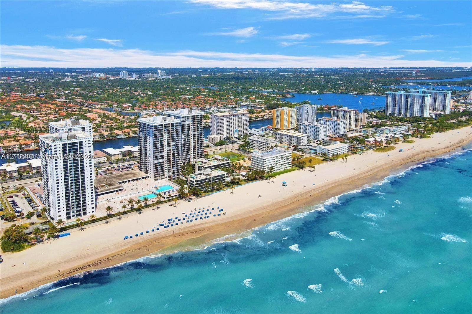 Coastal setting with expansive beach frontage, vibrant blue ocean, and a cityscape backdrop featuring high-rise buildings and a substantial body of water
