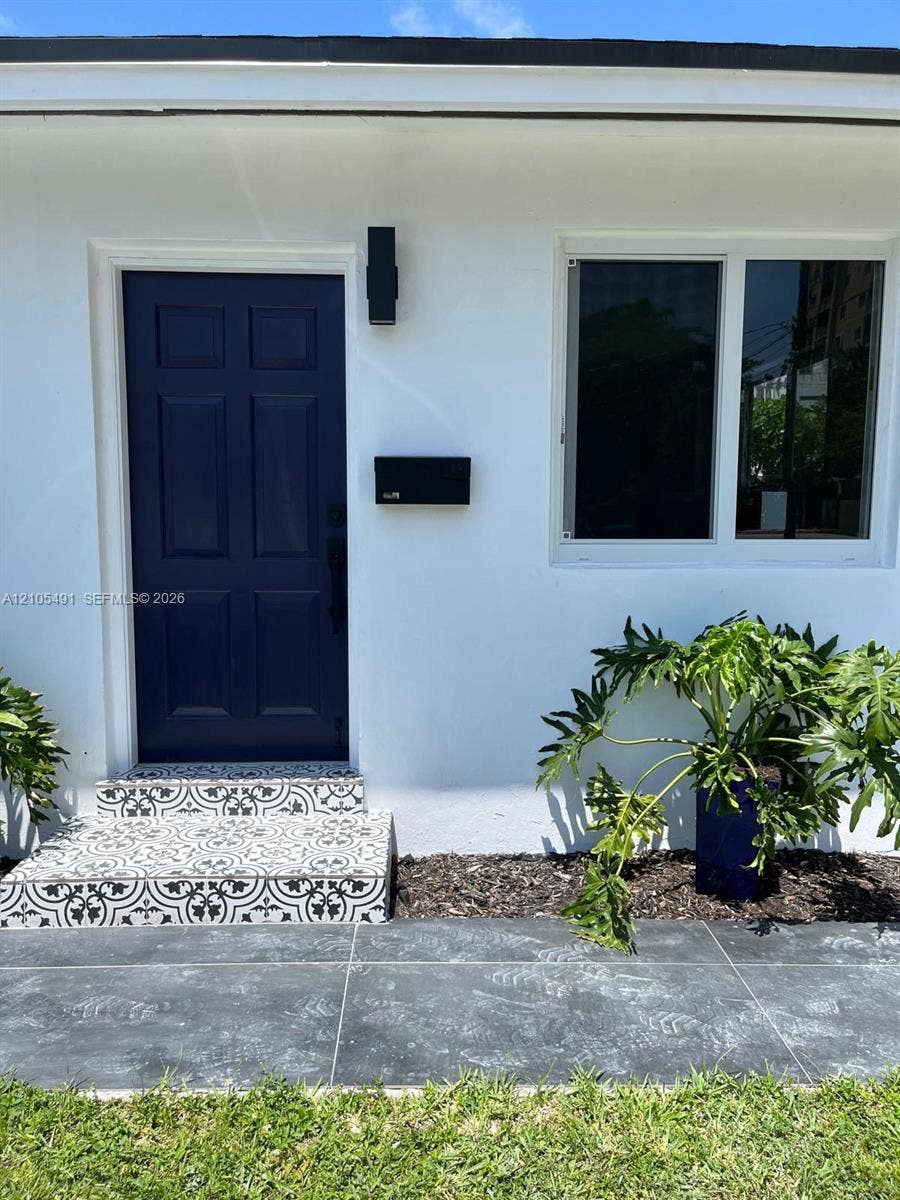 White exterior with contrasting navy paneled door, black wall-mounted light fixture, and patterned tile steps