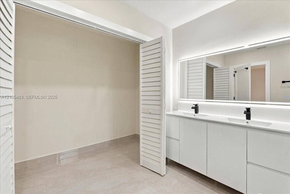 Double vanity with integrated sinks, white cabinetry, matte black hardware, and large illuminated vanity mirror, featuring louvered closet doors and tile flooring